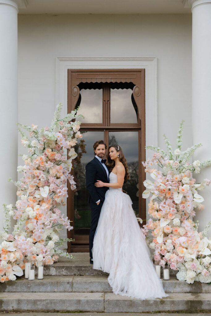 Bride and groom standing by a modern pastel floral asymmetric arch in a luxury styled shoot.